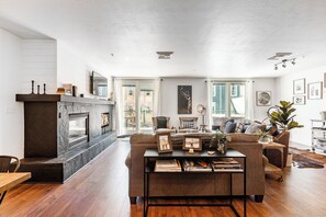A modern living room with a fireplace, a sofa, a coffee table with books, and a view of the outdoors through large windows.