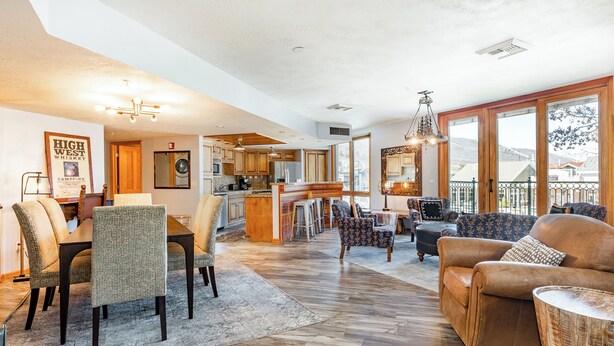 A spacious living area with a dining table, comfortable seating, a kitchen in the background, and large windows offering a view.