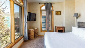 A hotel room with a large bed, a TV mounted on the wall, a wooden nightstand, and a window with curtains.