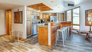 A modern kitchen with wooden cabinets, a large island with bar stools, and a view of the outdoors through large windows.
