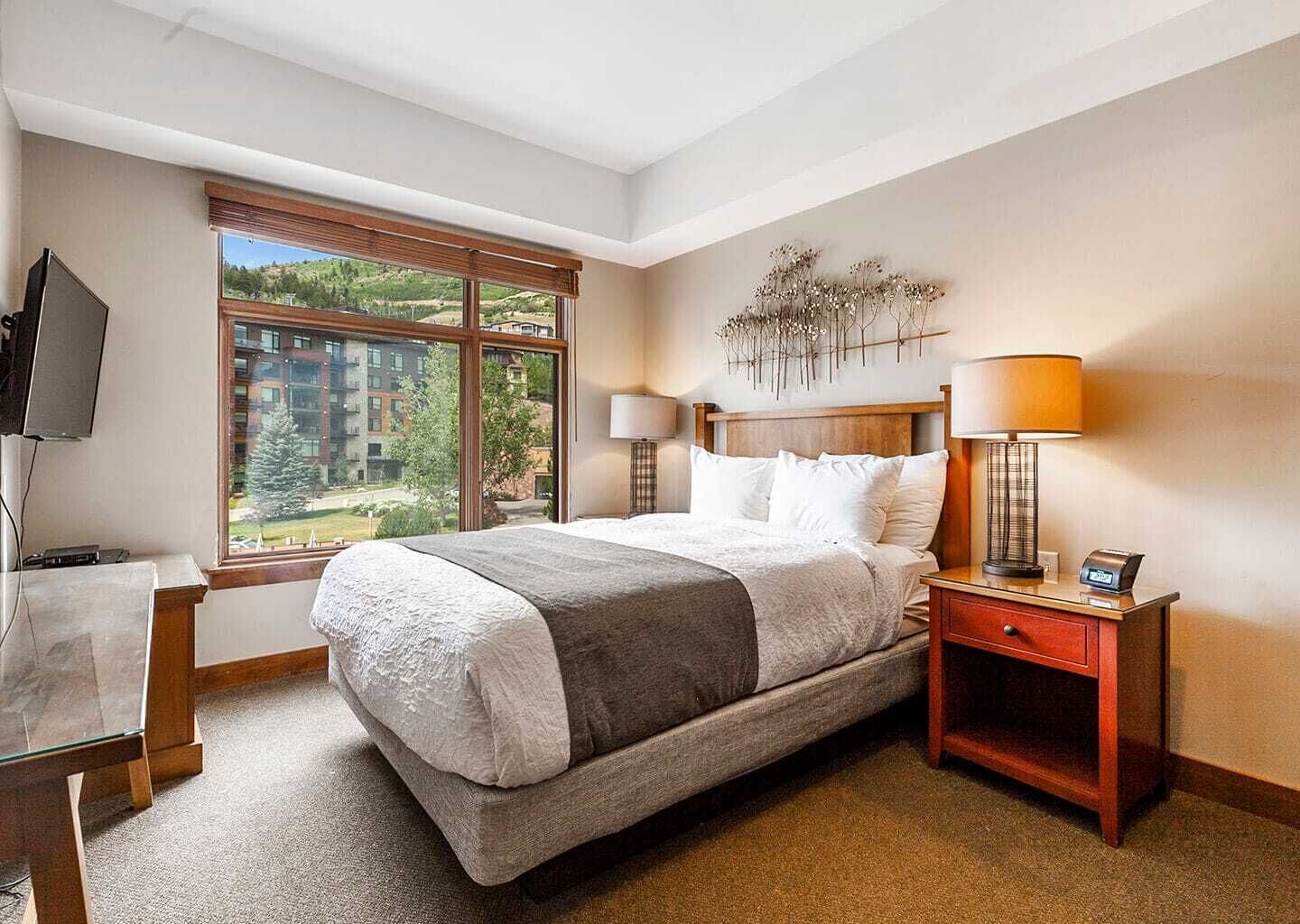 Une chambre d’hôtel avec un grand lit, un bureau avec une télévision, une fenêtre donnant sur des bâtiments et de la verdure, et un élément décoratif mural.