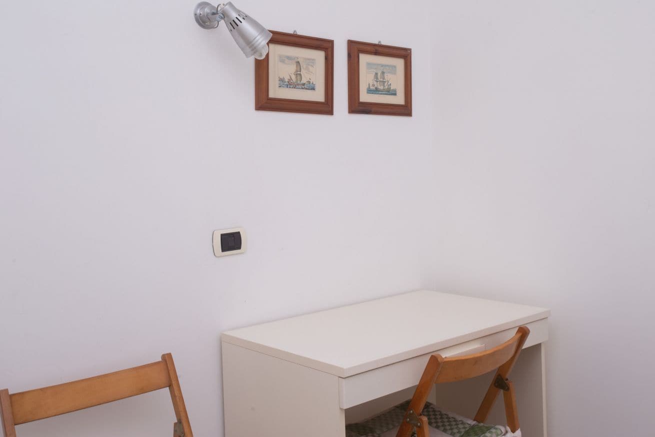 A room with a desk, two chairs, a wall-mounted lamp, and two framed pictures.