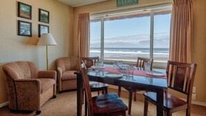 A room with a view of the ocean, a dining table set with glasses and plates, two armchairs, and a lamp. - Comfy Second Floor Beachfront Condo - Sleeps 4 - Pool and Sauna! (Lincoln City)