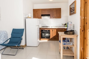 A compact kitchen with wooden cabinets, a white refrigerator, a blue chair, and a small wooden table with a frying pan.