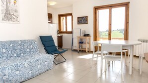 A living room with a patterned sofa, a blue chair, a white dining table with chairs, and a view through large windows. - M285 - Marcelli, comfortable three-room apartment with terrace (Marcelli)