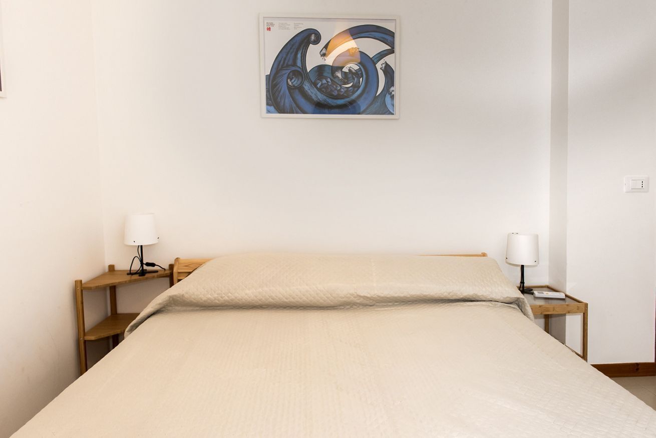 A bed with a beige cover, two bedside tables with lamps, and a wall-mounted abstract painting.