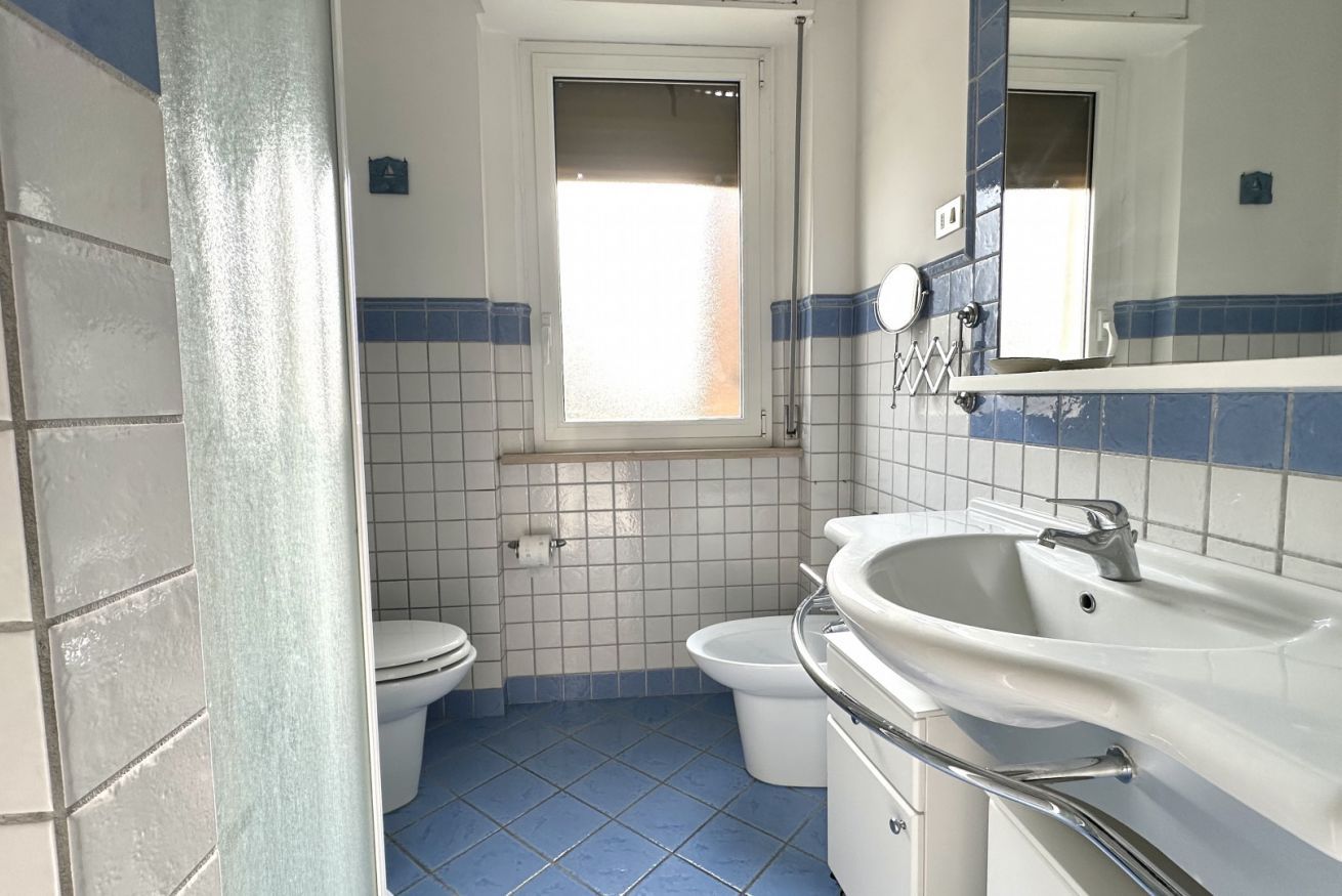 A bathroom with a toilet, bidet, and sink, tiled in blue and white, with a window above the toilet.