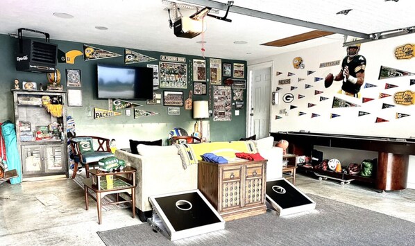 A room with a large TV mounted on the wall, a couch with a game table, and various sports memorabilia and flags on the walls.