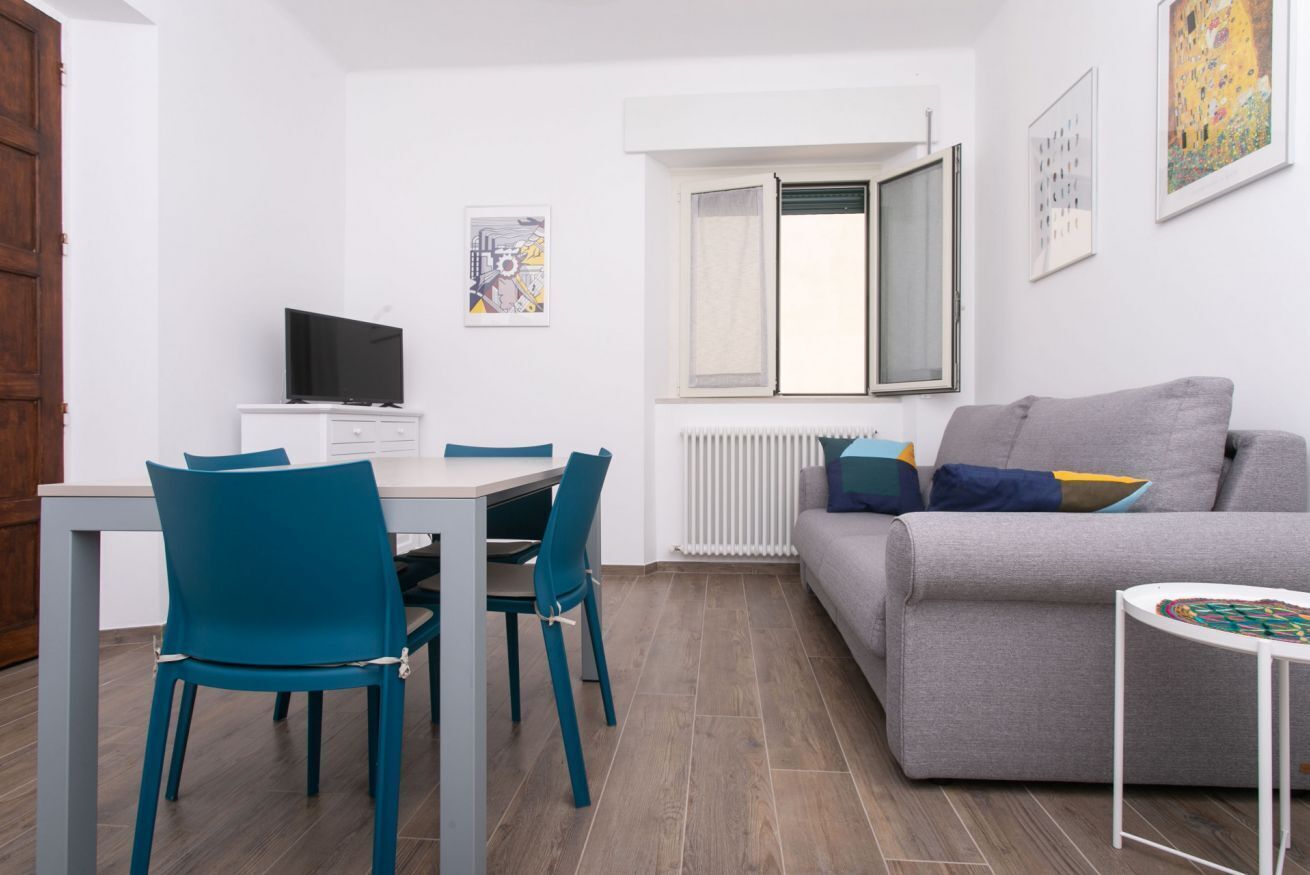 A modern living room with a grey sofa, a dining table with chairs, a TV, and a window with blinds.