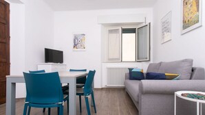 Een moderne woonkamer met een grijze bank, een eettafel met stoelen, een televisie en een raam met zonwering.