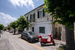 A street with a parked car and a red scooter, a building with a balcony, and trees along the sidewalk.