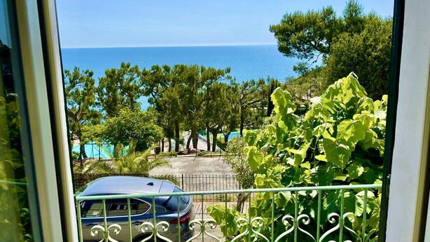 Een balkon met uitzicht op zee, een geparkeerde auto en groen.