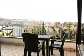 Un balcón con mesa y sillas, barandilla metálica y vista a un paisaje montañoso.