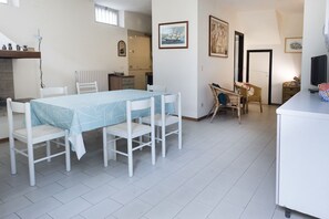 A dining area with a table and chairs, a kitchen in the background, and a television on a stand.