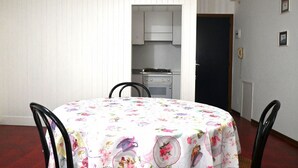 Un comedor con una mesa redonda cubierta con mantel floral, sillas negras y una cocina al fondo.