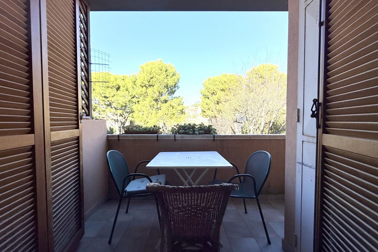 Balcón abierto con mesa y sillas, vista a árboles y edificios, y una puerta con persiana cerrada.