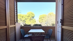 Een open balkon met een tafel en stoelen, uitzicht op bomen en gebouwen, en een gesloten luik.