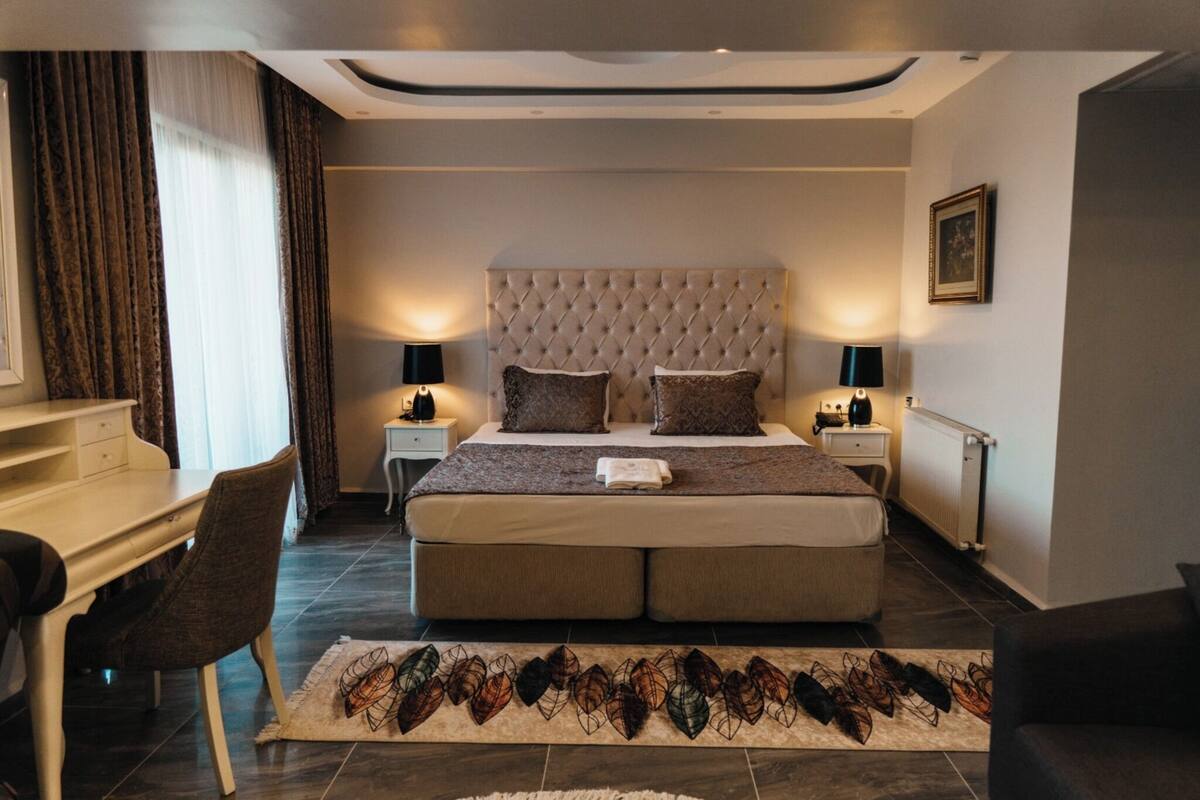 A bedroom with a tufted headboard, a desk, a chair, a bedside table with lamps, and a radiator.