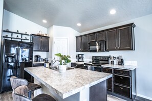 A modern kitchen with dark cabinets, a stainless steel refrigerator, a built-in oven, and a marble island. - Beautiful Oasis with a splash of Glam (Kuna)