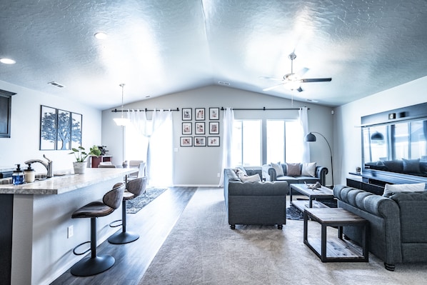 A spacious living area with a large sofa, a coffee table, and a TV. There is a kitchen area with a bar and stools. The room has a high ceiling with a fan and large windows. - Beautiful Oasis with a splash of Glam (Kuna)