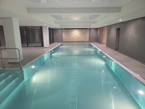 Una piscina interior larga y estrecha con suelo texturizado, rodeada de paredes de apariencia pétrea e iluminación empotrada.