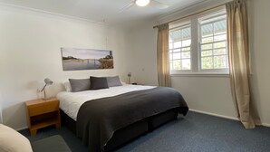 A bedroom with a bed, bedside tables, a painting, a window with curtains, and a ceiling fan. - Allamanda House, 3 Willow Street (Crescent Head)