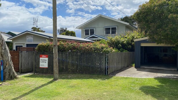 A residential property with a fenced yard, a garage, and a house with a sign indicating holiday letting. - Allamanda House, 3 Willow Street (Crescent Head)
