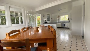 A spacious dining area with a wooden table and chairs, a kitchen with white cabinets, and a tiled floor. - Allamanda House, 3 Willow Street (Crescent Head)