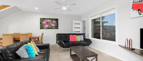 A modern living room with a black leather sofa, a wooden coffee table, a dining area with a wooden table and chairs, a large window with a view, and a ceiling fan.