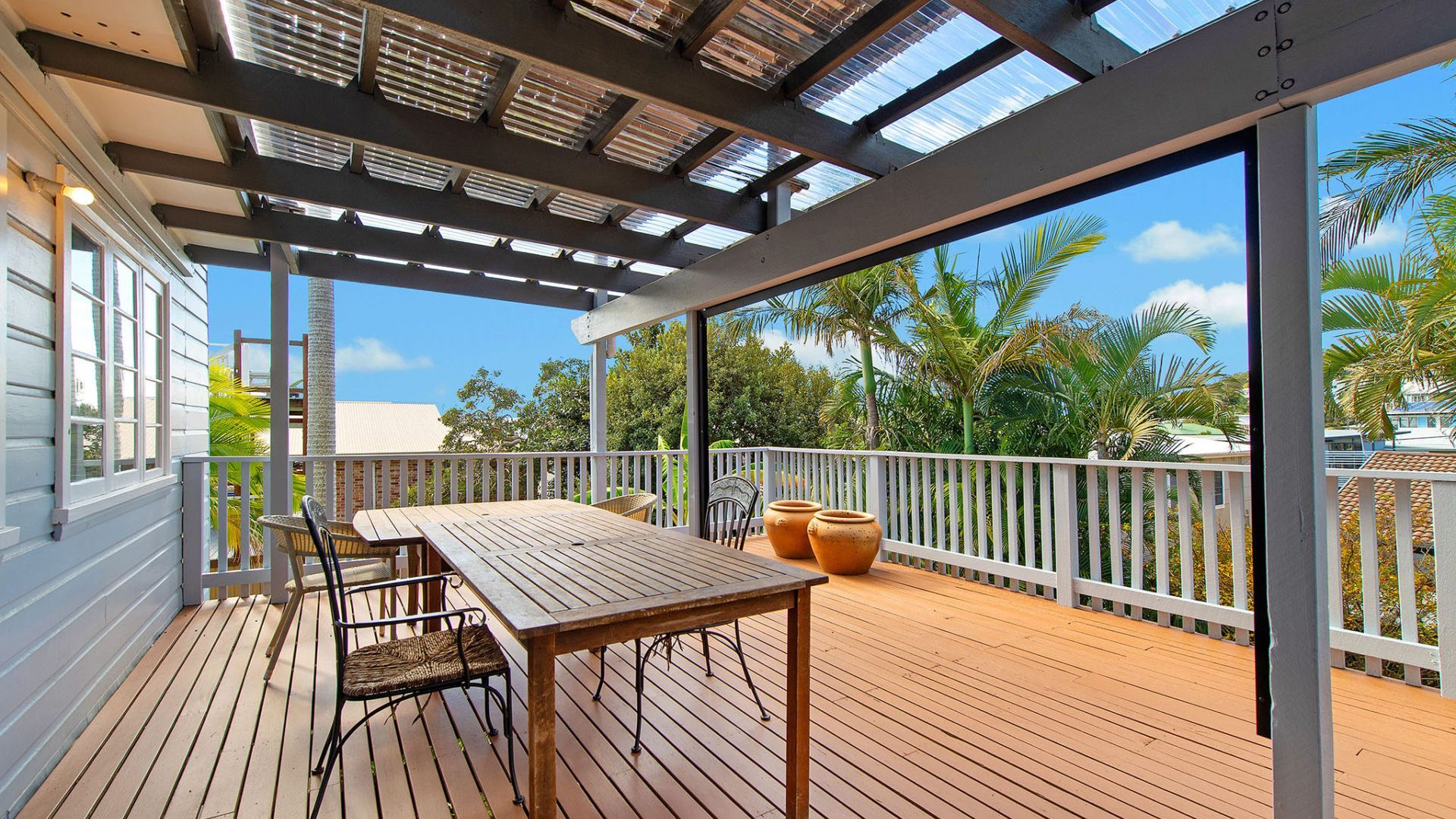 An outdoor deck with a wooden table and chairs, a pergola with a roof, and a view of palm trees and a building in the background.