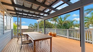 An outdoor deck with a wooden table and chairs, a pergola with a roof, and a view of palm trees and a building in the background. - Berry, 16 East Street (Crescent Head)