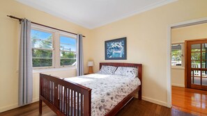 A bedroom with a wooden bed, a window with curtains, a painting on the wall, and a door leading to an outdoor area.