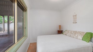 A bedroom with a bed, a small bedside table, a hanging macrame decoration, and a window with a view of greenery.