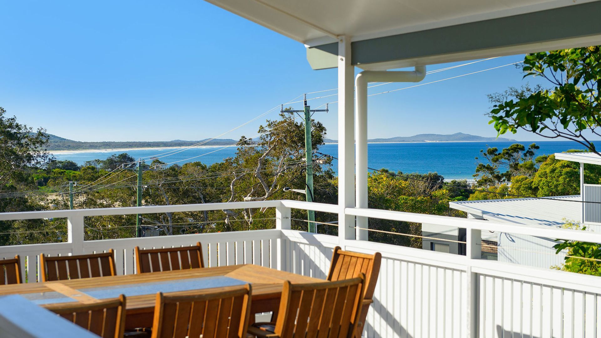 Un balcon avec une table et des chaises, donnant sur un paysage côtier avec des arbres et une vue sur la mer.
