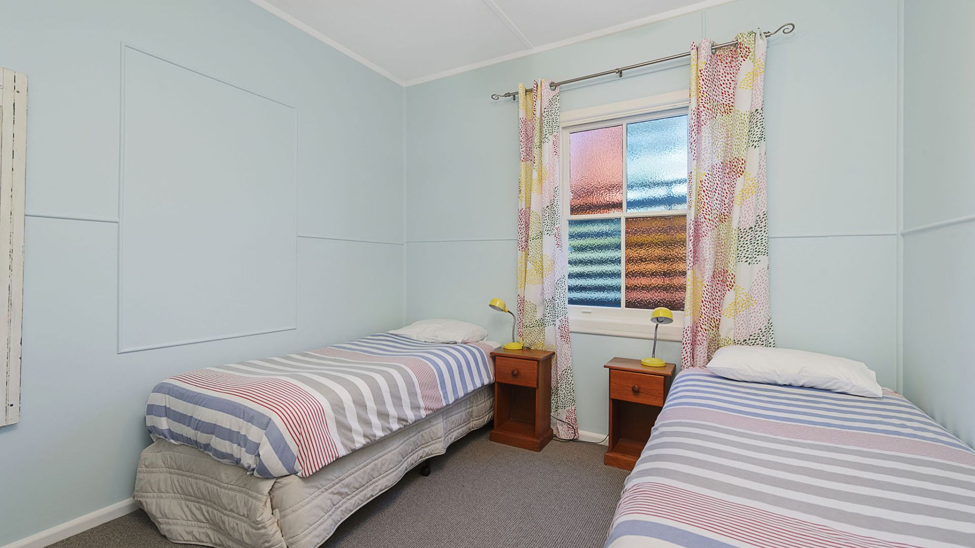 Une chambre avec deux lits simples, une fenêtre avec des rideaux à motifs, une table de chevet avec une lampe et une étagère fixée au mur.