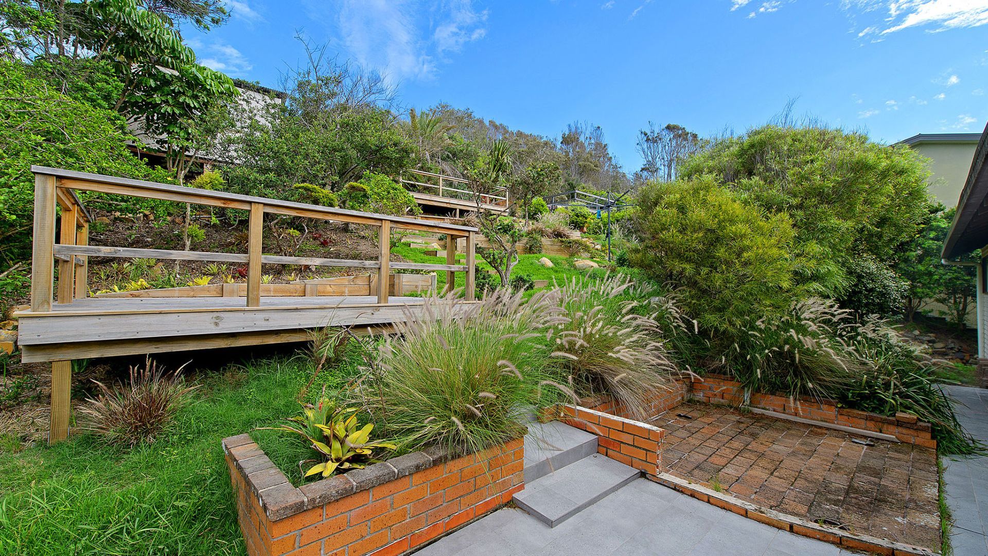 A wooden bridge over a garden with brick planters and a variety of plants.
