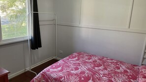 A small, single-bed room with a window, a metal wardrobe, and a red patterned bedspread.