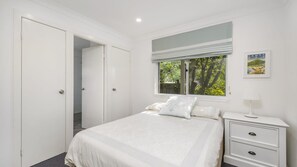 A bedroom with a bed, a nightstand, a window with a view of trees, and a door leading to another room. - Mirador, 1 Stewart Street (Crescent Head)