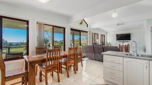 A spacious living area with a dining table and chairs, a sofa, a kitchen with a sink and cabinets, and large windows offering a view of the outdoors.