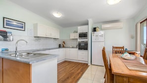 A compact kitchen with white cabinets, a built-in oven, and a dining area with a wooden table and chairs. - Heathers Seaside Apartments 1, 10 Rankine Street (Crescent Head)