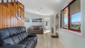A living room with a leather sofa, a wooden wall with nautical decorations, a view through a window, and a tiled floor. - Heathers Seaside Apartments 1, 10 Rankine Street (Crescent Head)