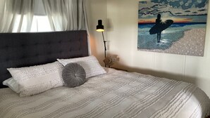 Een net opgemaakt bed met een gestreept dekbed en kussens, een bedlampje en een wanddecoratie van een surfer.