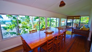 A spacious dining area with a large wooden table and chairs, large windows offering a view of lush greenery and a body of water. - Rosemaree, 24 East Street (Crescent Head)