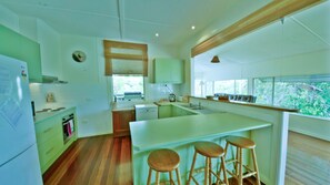 A kitchen with a central island, wooden stools, and a view of greenery through large windows.
