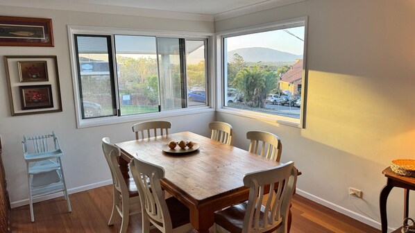 A dining area with a wooden table and six chairs, large windows with a view of the outside, and two framed pictures on the wall.