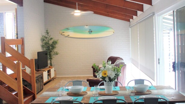 Un comedor con una mesa de madera dispuesta para cuatro, una tabla de surf en la pared y una planta en la esquina.