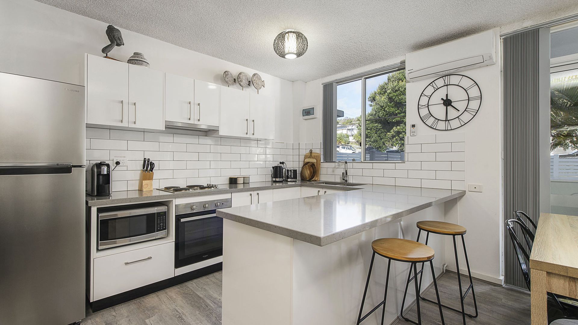 A modern kitchen with white cabinetry, stainless steel appliances, a large island with stools, and a wall clock.