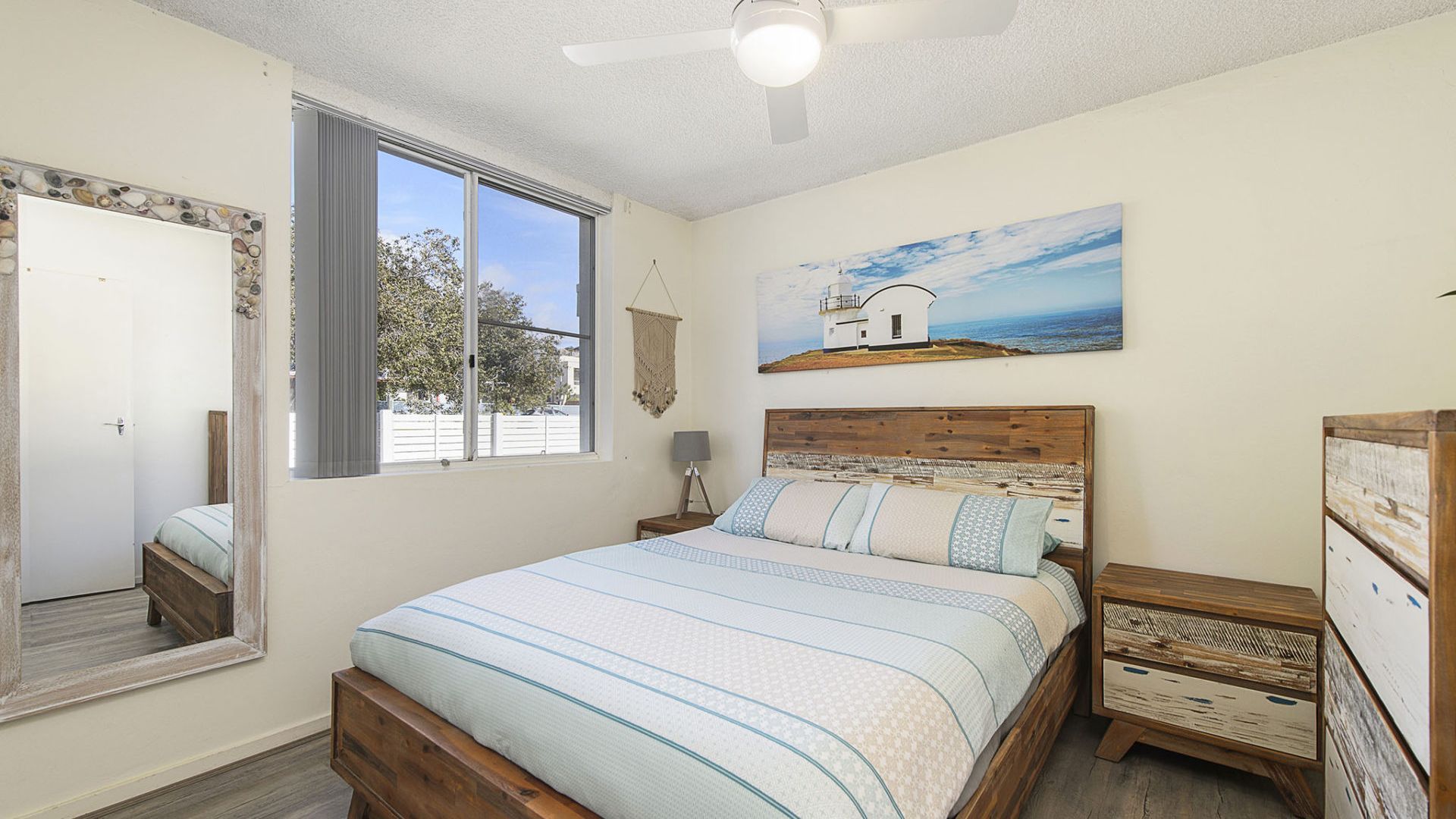 Une chambre à coucher comprenant un lit, une table de chevet, une commode, un ventilateur de plafond, une fenêtre et un tableau accroché au mur.