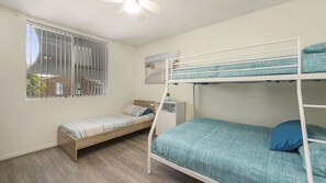 Une chambre avec des lits superposés, un lit simple, une fenêtre avec des stores, un ventilateur de plafond et une petite table avec une plante.