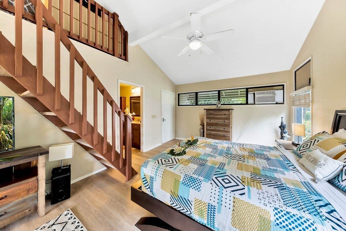 A bedroom with a large bed, a wooden staircase, a ceiling fan, and a view of the bathroom.